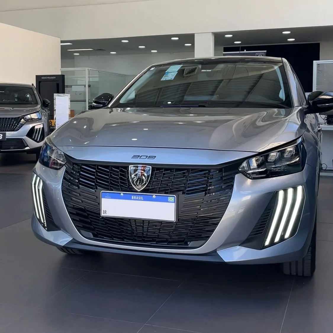 Carro Peugeot 208 prata exibido em uma concessionária, destaque para o design moderno, faróis de LED e detalhes do interior ao fundo.