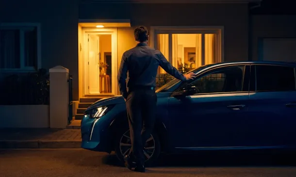 Pessoa de terno parando ao lado de um carro na rua à noite, diante de uma casa iluminada com luz quente na entrada.