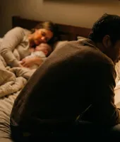 Cena de uma mulher triste segurando um bebê na cama, enquanto um homem triste sentado ao lado, em um quarto com iluminação suave e ambiente acolhedor.