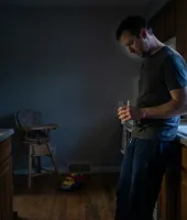 Homem pensativo na cozinha, segurando um copo de água, ambiente com iluminação suave da manhã ou fim de tarde, mostrando uma rotina diária.